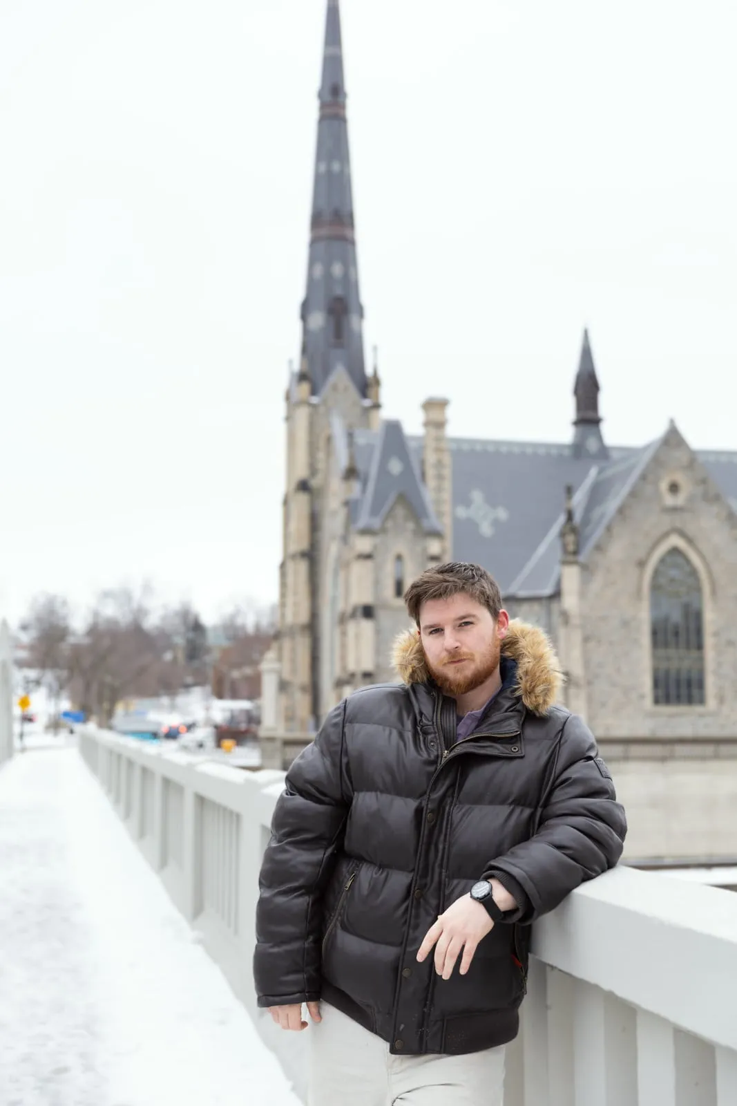 A man in a winter jacket on a bridge, a church spire behind him
